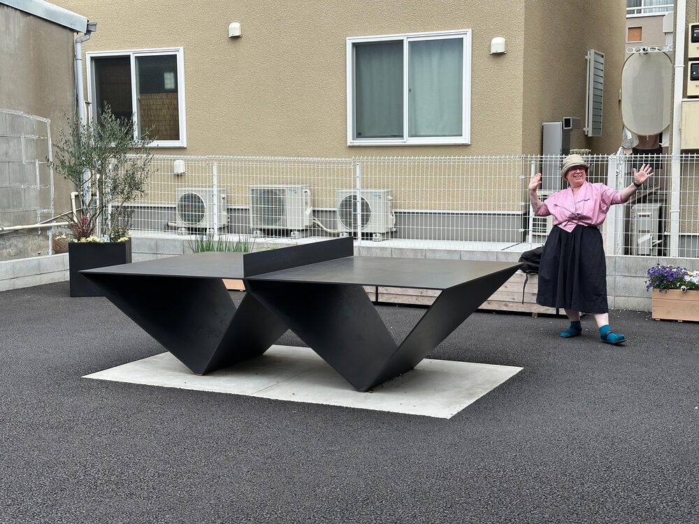 Nadja standing with arms wide at a sculptural outdoor table-tennis table in Japan